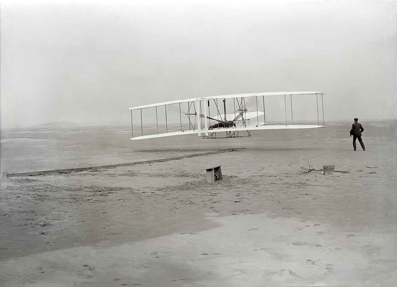 Photographie noir et blanc du Wright Flyer décollant au-dessus du sable, Wilbur Wright courant à côté de l'aile.